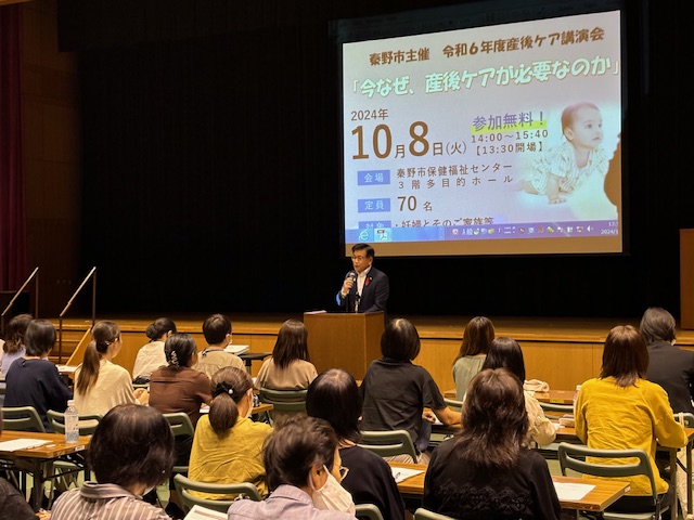 令和6年度産後ケア講演会で、演台に立つ市長が「今なぜ、産後ケアが必要なのか」というスライドを背景に聴衆へ向けて話しているイベント風景の写真