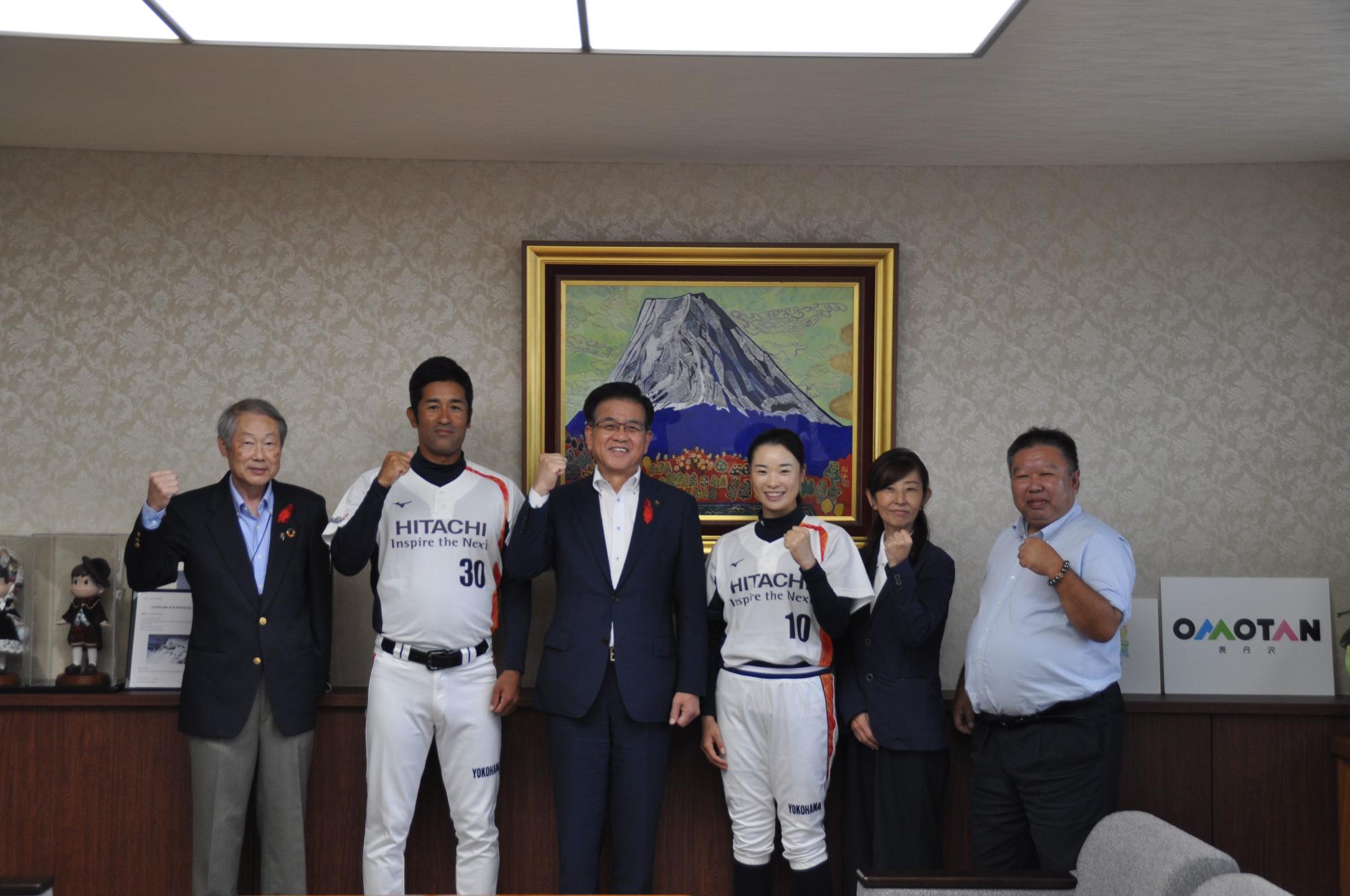 富士山の絵が飾られた部屋で、スポーツユニフォームを着た男女2人と市長を含めたスーツ姿の男女4人が並び、全員がこぶしを上げて記念撮影している写真
