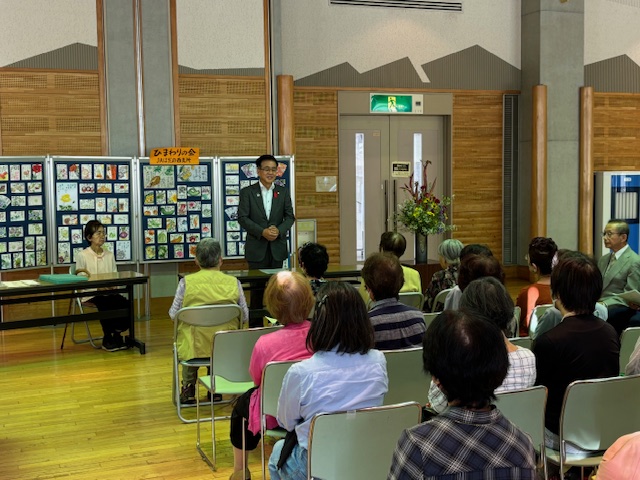 屋内で行われているイベントで、作品が展示されている会場に集まった人々が、マイクを持って挨拶する市長に注目している様子の写真