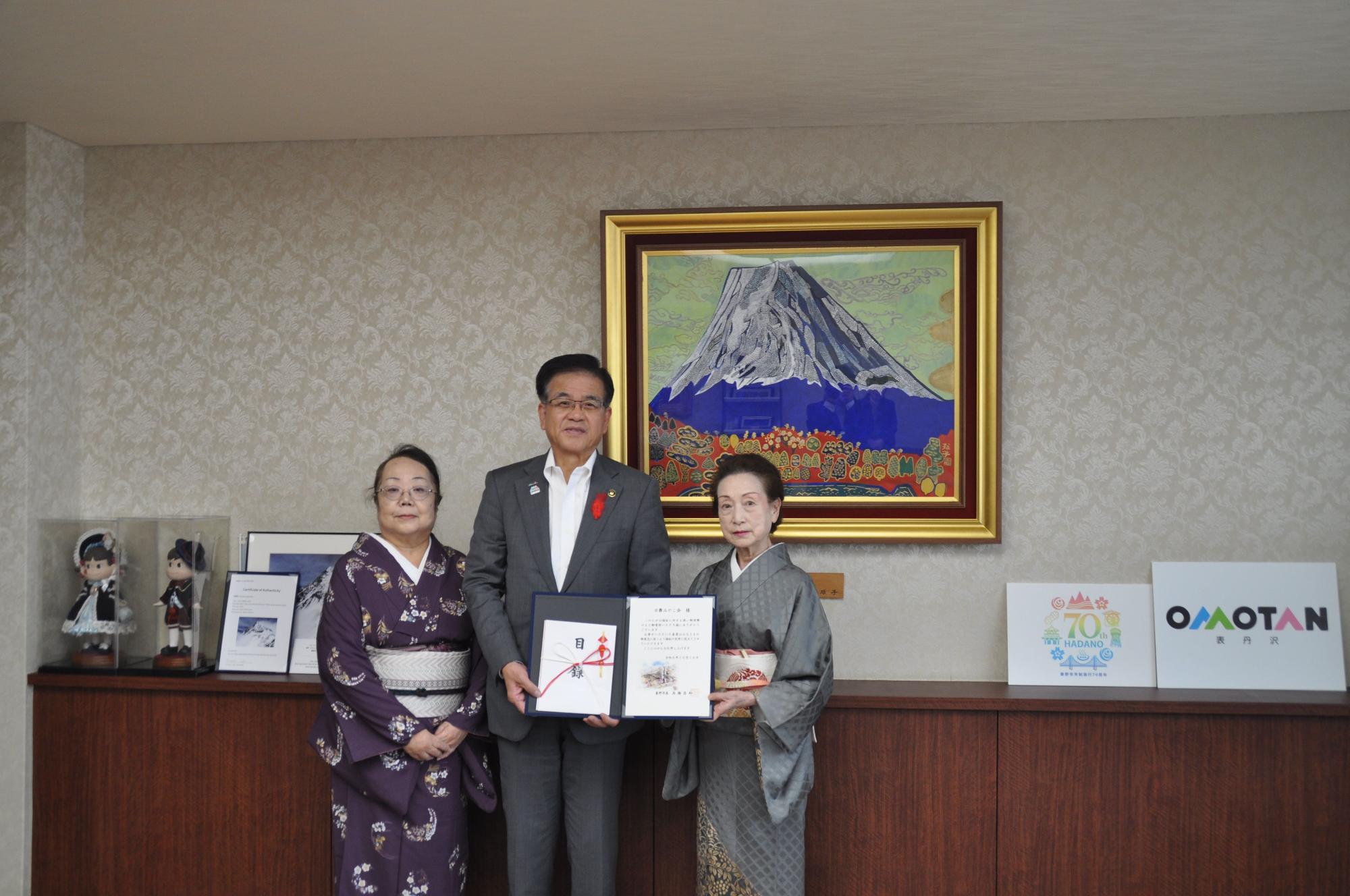 壁に富士山の絵が飾られた部屋で、市長を中心に着物姿の二人の女性と一緒に目録などを手に記念撮影をしている写真