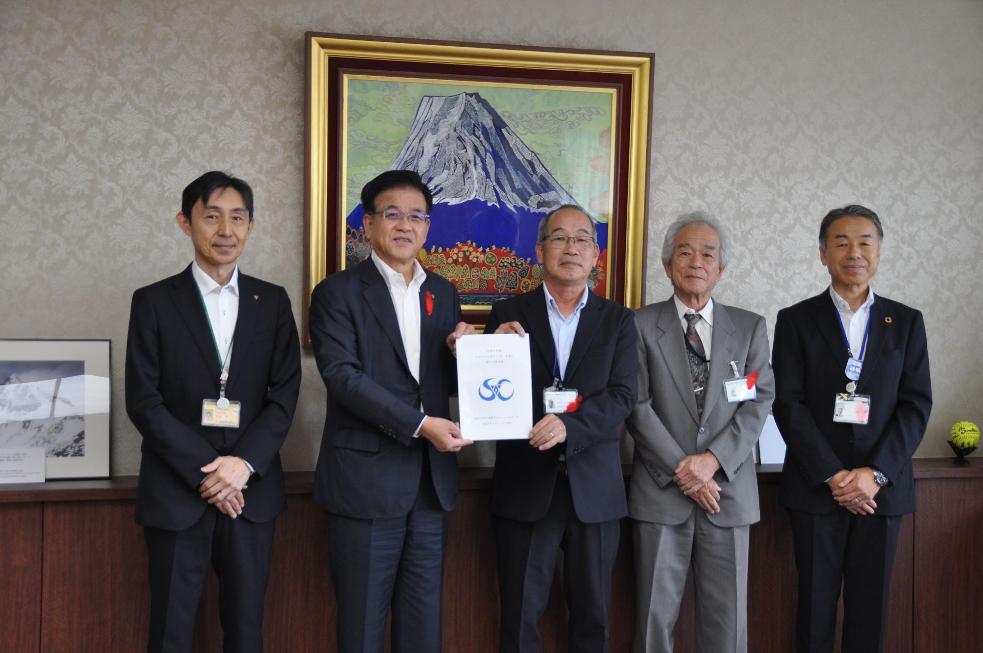 富士山の絵が飾られた部屋で、5人の男性が並んでおり、中央の市長と男性がロゴの入った要望書のようなものを一緒に持っている記念写真