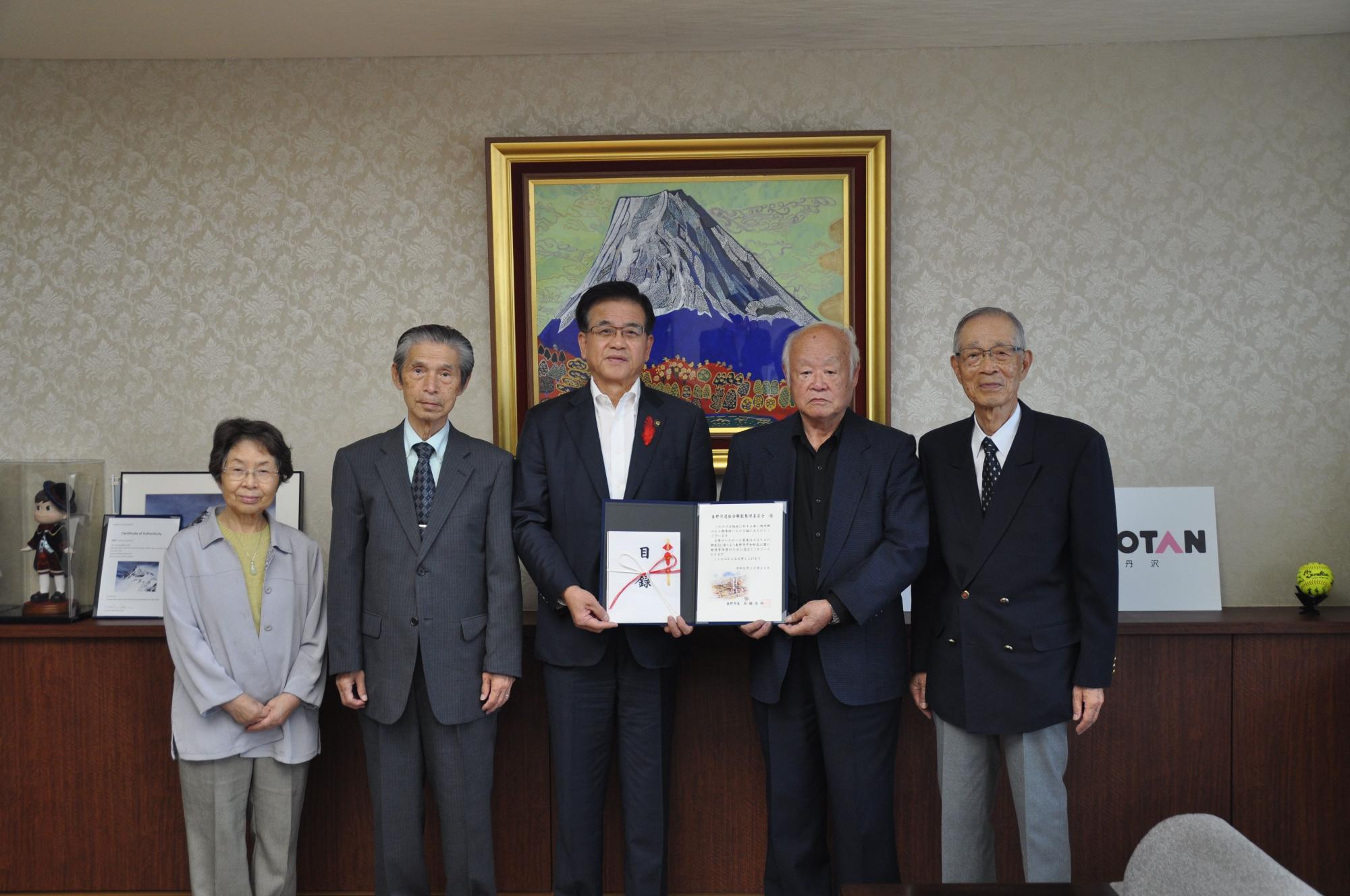 5人の男女が部屋に並んでおり、そのうちの市長と関係者の男性が目録などを持って微笑んでいる集合写真