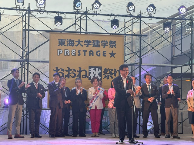 「東海大学建学祭」「おおね秋祭り」と書かれた横断幕が掲げられた舞台で、マイクを持って話す市長を、複数の人々が囲んでいるイベント風景の写真