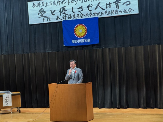 木製の演台に立つ市長が、マイクを手に聴衆へ向けて話をしている室内写真