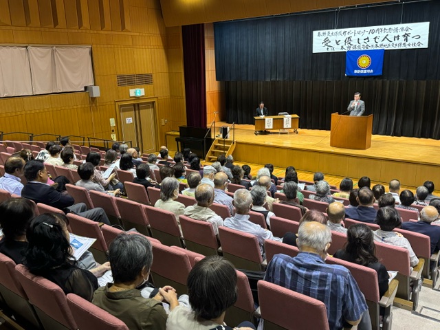 「愛と優しさで人は育つ」と書かれた横断幕が掲げられた講堂の舞台で、演台に立つ男性が大勢の聴衆に向かって話している講演会の写真