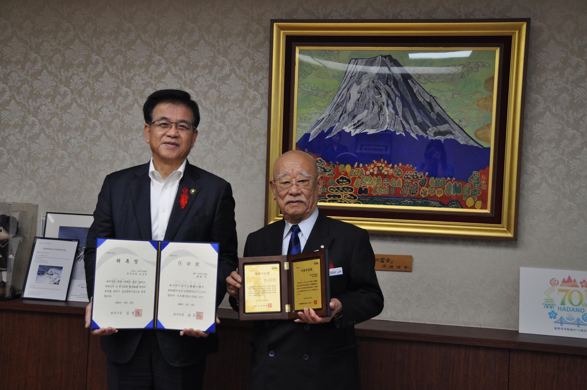 壁に富士山の絵が飾られた部屋で、市長と福森会長がそれぞれ賞状と盾を持って微笑んで、記念撮影をしている様子の写真