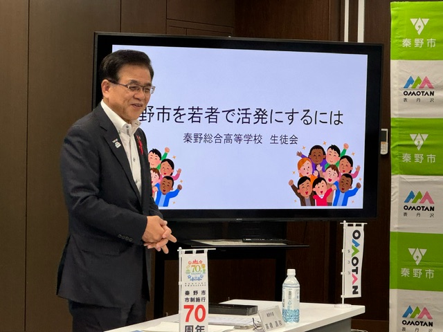 市長が笑顔で話しており、背景にあるスクリーンには「秦野市を若者で活発にするには」というタイトルが映し出されている写真