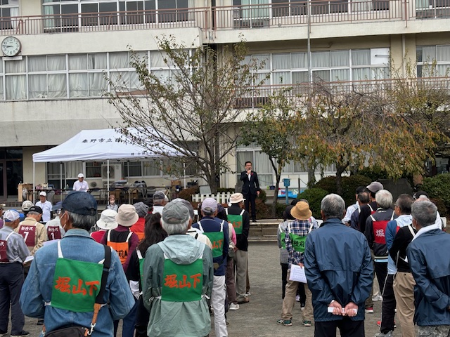 ビブスを着用した参加者たちが、学校の校庭でマイクを手に挨拶する男性の方を向いて整列している屋外イベントの写真