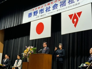 背景には日の丸と秦野市のマークが入った旗が掲げられている壇上の演台でマイクに向かって話す市長と手話通訳者が立っている写真