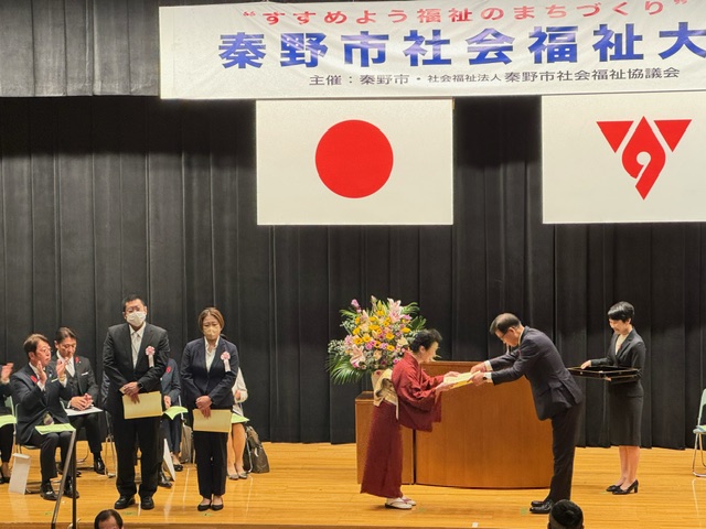 表彰式で着物姿の女性が市長から表彰状を受け取っており、その傍に胸章をつけた男女が立っている壇上の写真