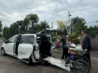 白い介護車両の後部から車いす用のスロープが伸びており、二人の男性が車両の横に立ったまま話している屋外での写真