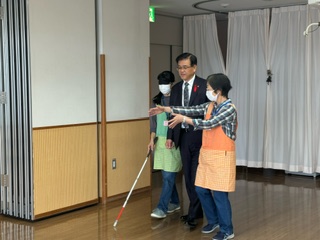 アイマスクをした人が白杖を使いながら、付き添い役の市長と関係者の女性に誘導されて室内を歩いている、視覚障害体験をしている様子の写真