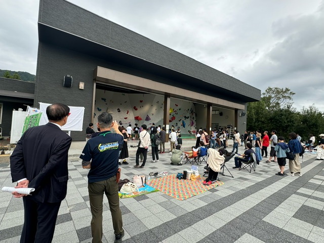 建物前の広場で開催されている屋外ボルダリングイベントで、多くの人がカラフルなホールドが取り付けられた壁を見上げている様子の写真