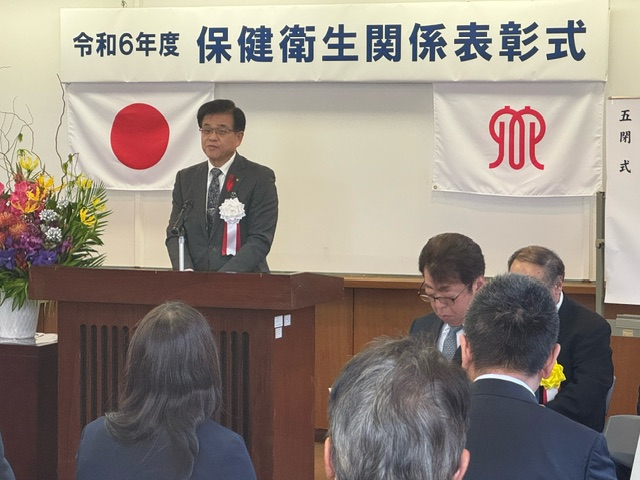 令和6年度の保健衛生関係表彰式で、日の丸と赤いシンボルマークの旗が掲げられた演壇で挨拶する市長の写真