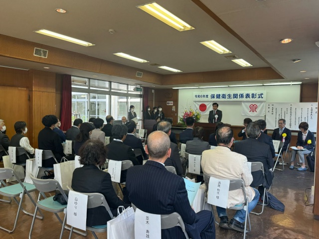 令和6年度の保健衛生関係表彰式会場で、大勢の参加者が椅子に座って演壇の様子を見守っているイベント風景の写真