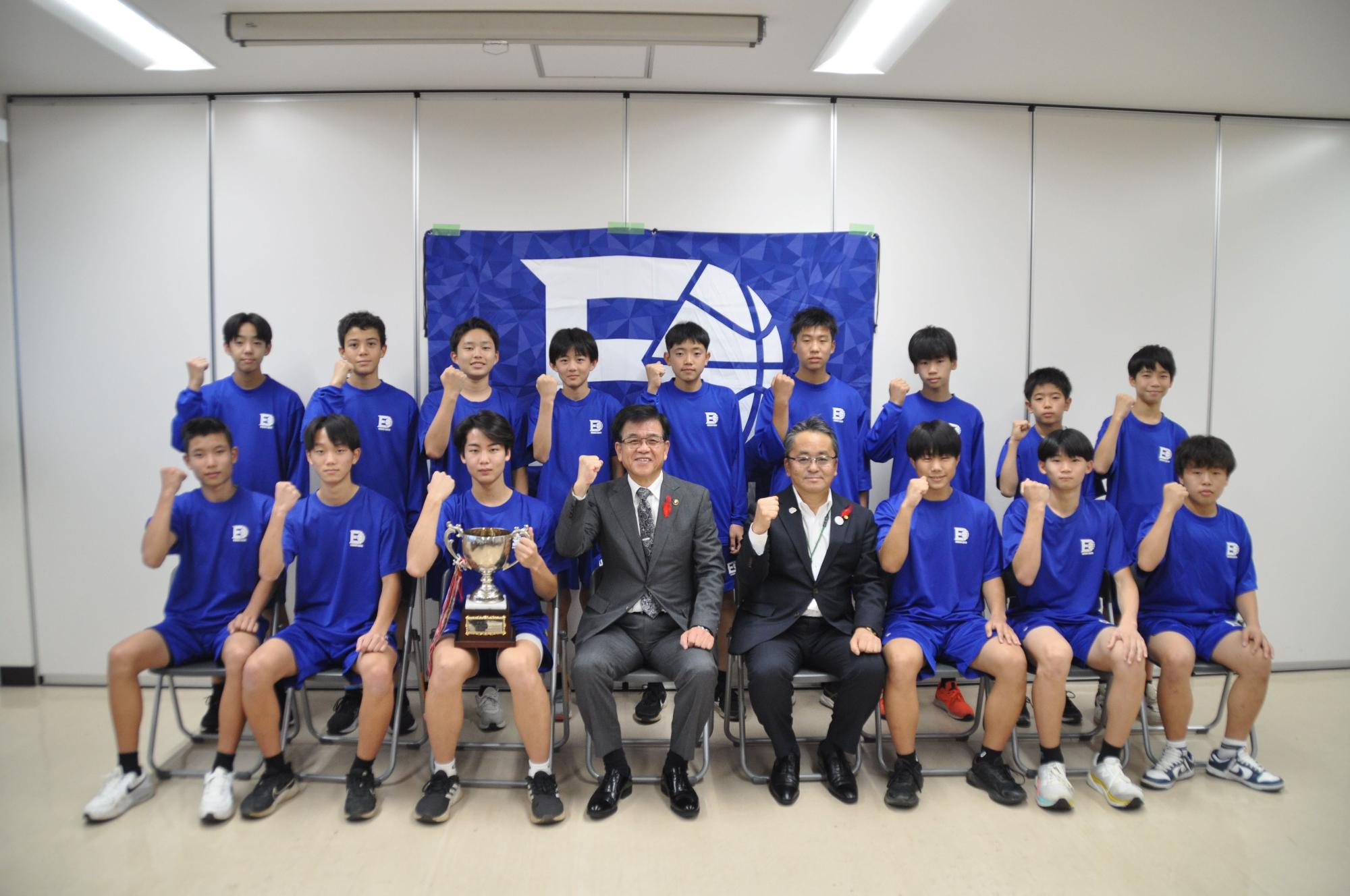ブルーのユニフォームを着た男子バスケットボールチームのメンバーたちが、前列に座る市長と関係者二人が一緒にトロフィーを囲んで写っている集合写真