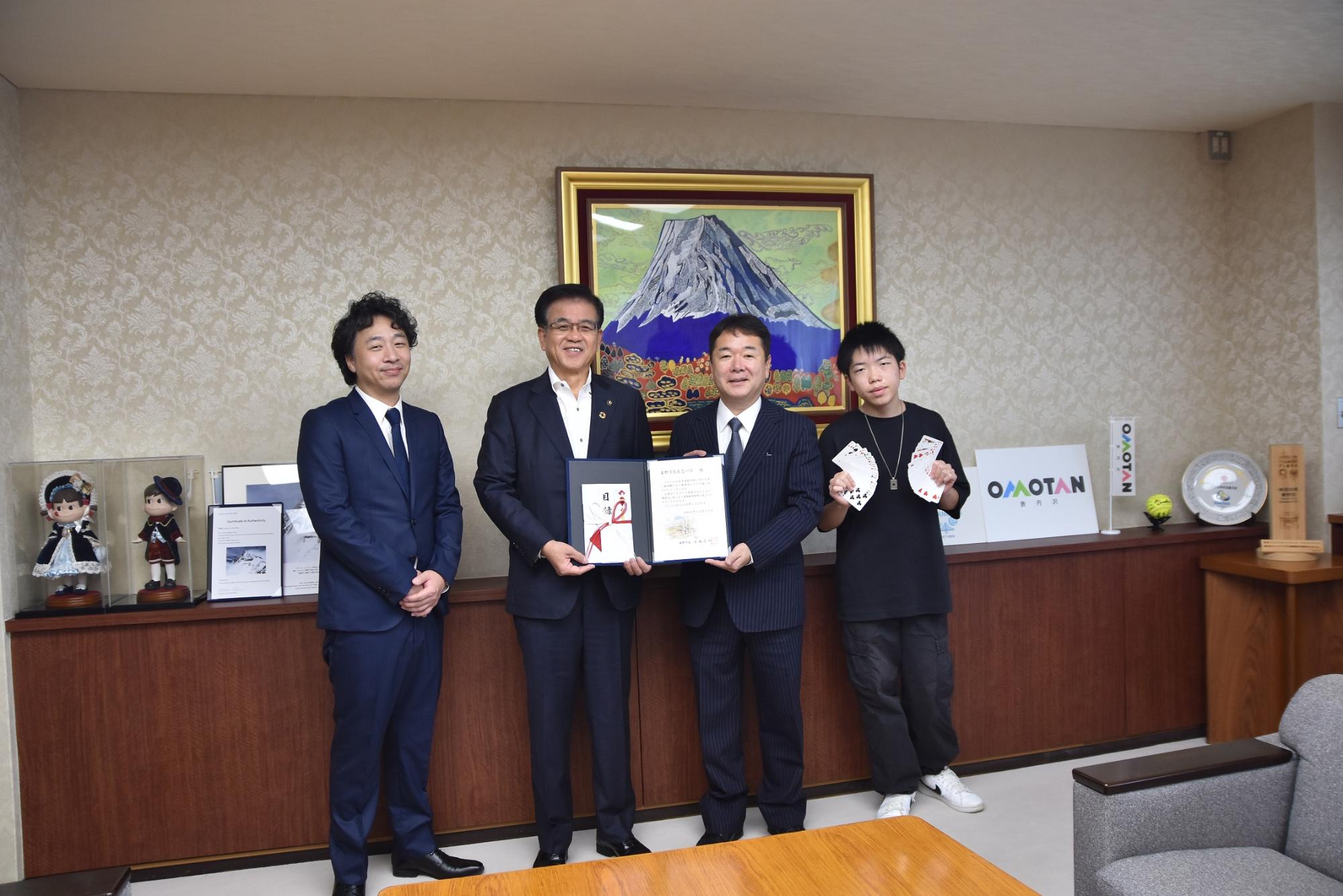 四人の男性が部屋の中で記念撮影をしており、そのうち市長と関係者二人が表彰状を持ち、一人の男性がトランプを手にしている写真