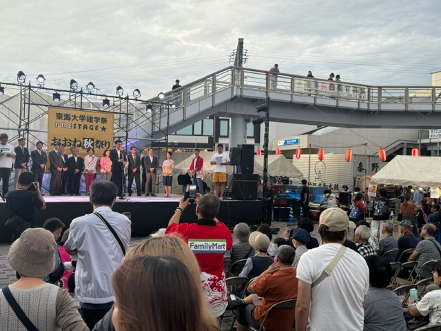 「東海大学建学祭」「おおね秋祭り」と書かれた横断幕がある屋外のステージで、大勢の観客を前に複数の人物が並んでいるイベント風景の写真