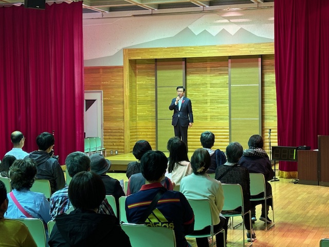 茶色の木目調の壁と赤いカーテンの舞台上で、市長がマイクを手に、椅子に座っている大勢の聴衆に向かって話している写真