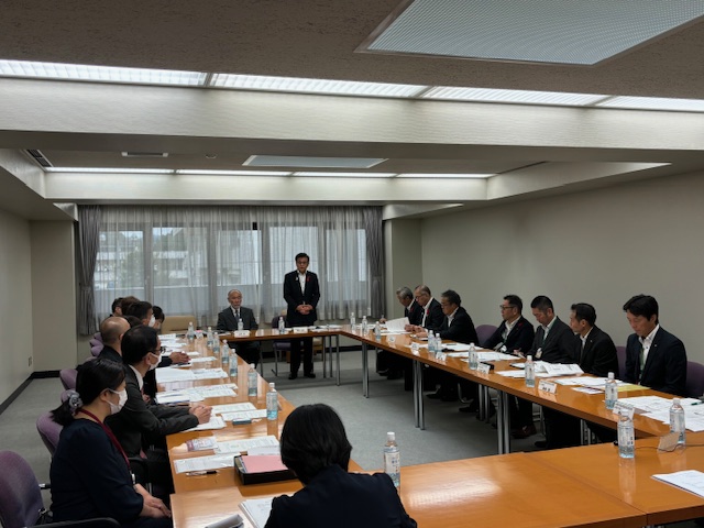 広々とした会議室で、大きなロの字型のテーブルを囲んで複数の人々が着席しており、中央でスーツ姿の男性が立って話をしている会議風景の写真