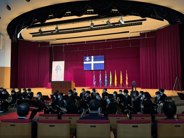 色とりどりの旗が立てられたステージを背に、楽器を持った大勢の学生たちが、客席を向いて座っている様子の写真