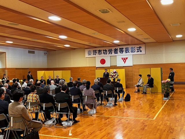 「秦野市功労者等表彰式」の横断幕と日本国旗、市章が掲げられた壁の前に金屏風が設置され、その前の演台に立った高橋市長が、着席した大勢の出席者に向かって話をしている写真