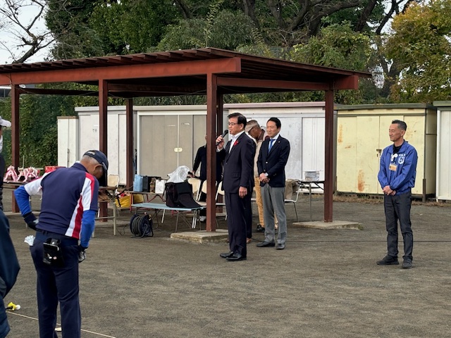 屋外の屋根付きベンチの横に立った高橋市長が、関係者や参加者が並ぶ中マイクで話をしている写真