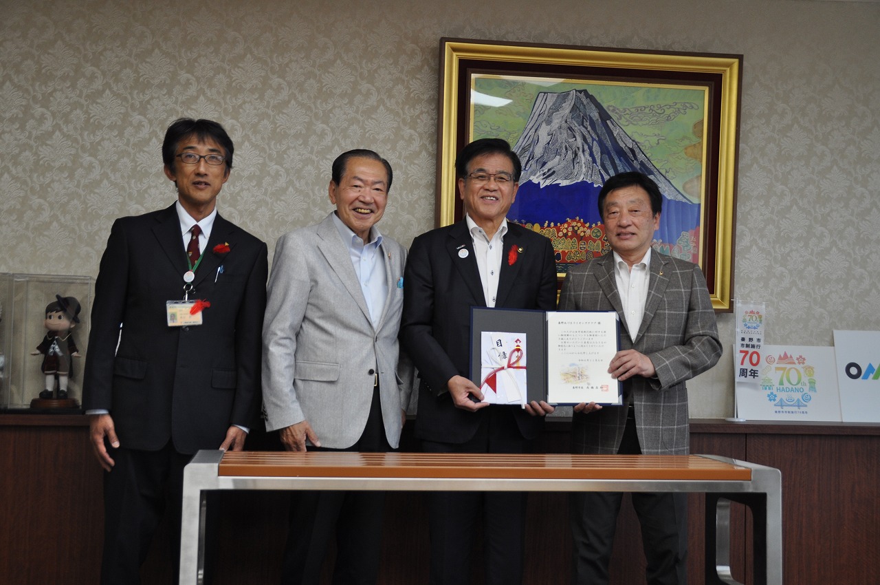 山の絵画が掛けられた壁を背に、高橋市長と尾谷会長ほか2人の男性が並び、手前に寄贈されたベンチを置いて撮影した記念写真