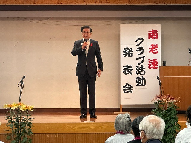 「南老連クラブ活動発表会」の立て看板が置かれた舞台上で、マイクを持った高橋市長が客席に向かって話をしている写真