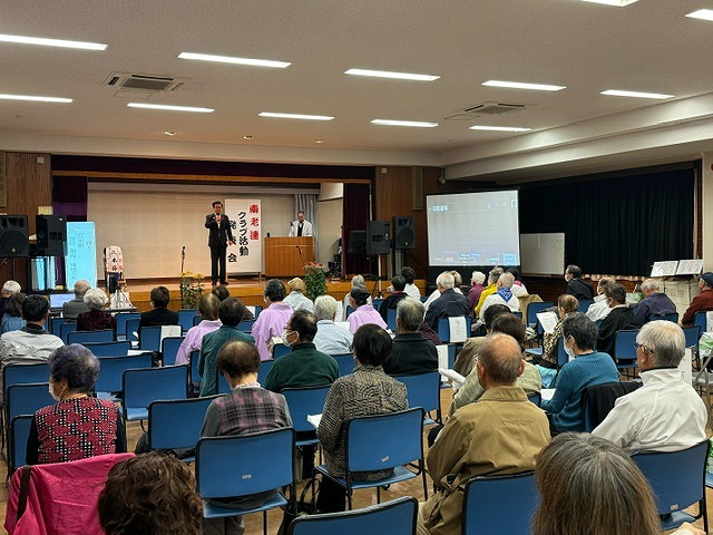 会場いっぱいに並んだ椅子にたくさんの出席者が座り、奥のステージに上がった市長が話をしている様子の写真