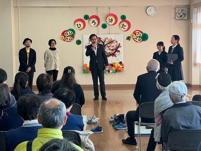 「カムカムひろはたフェスタ」の文字が装飾された壁の前に2人の男女と2女子高校生が立ち、中央に立った高橋市長が席に座った参加者たちに向かって話をしている写真