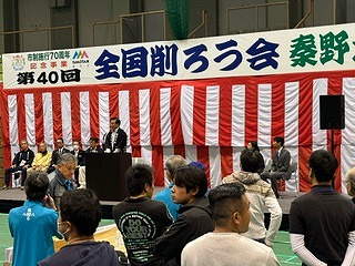 「第40回全国削ろう会」の横断幕が設置された紅白幕の前に複数の関係者が座り、演台に立った市長が会場に向かって話をしている写真
