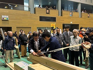 大勢の人々が見守る中、男性の指導を受けながら法被姿の市長が角材をカンナで削っている様子の写真