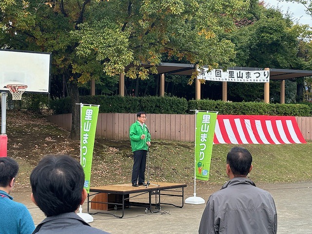 木々の茂る公園で、緑色のジャンパーを着た市長が、「里山まつり」と書かれた緑色ののぼりの間にあるステージに立って、出席者に向かってスピーチをしている写真