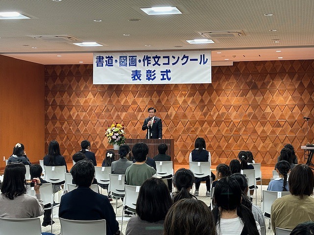 「書道・図画・作文コンクール表彰式」の横断幕が設置された、ひし形の木目調の壁を背に演台に立った高橋市長が、席に座ったたくさんの出席者たちに話をしている写真