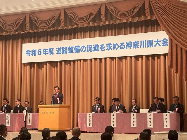 「令和6年度道路整備の促進を求める神奈川県大会」の横断幕が掲げられた壇上に設けた席に複数の関係者が座り、その前の演台に立った高橋市長が話を聞いている様子の写真