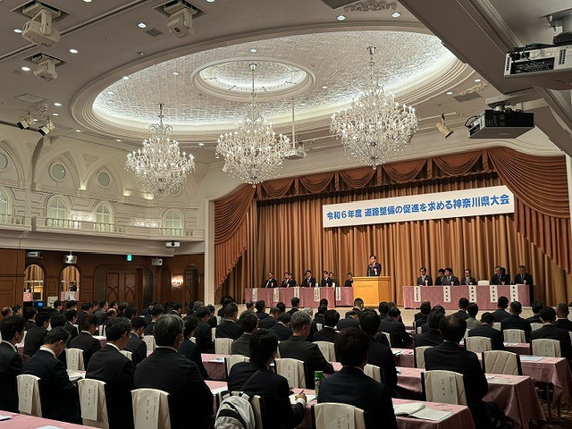 3つのシャンデリアが吊り下げられた広々としたホールの壇上に設けた席に複数の関係者が座る中、一人の男性が演台に立っており、客席には大勢の参加者たちが座っている様子の写真