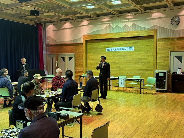 ホールに囲碁盤を乗せたテーブルが複数並び、参加者の男性たちが席に座る中、前に立った高橋市長がマイクで話をしている写真