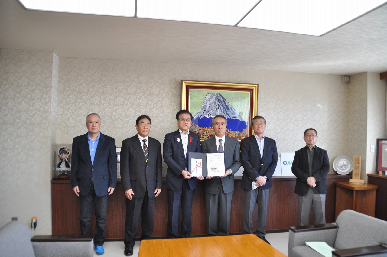 山の絵画が掛けられた壁を背に、目録を持った高橋市長と小室管理委員長を挟み、4人の男性が並んで立っている記念写真