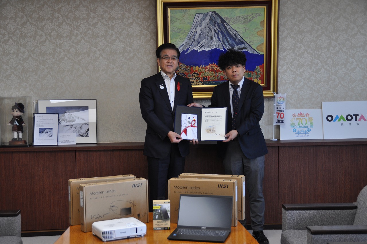 山の絵画が掛けられた壁を背に目録を持った高橋市長と久宗店長が並んで立ち、手前のテーブルに並んだノートパソコンなどの電子機器と一緒に写した記念写真