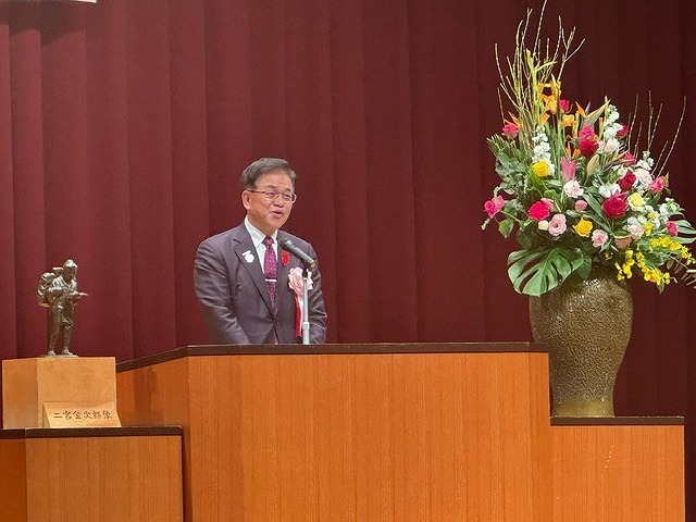 豪華な花と二宮金次郎の小さな像が飾られた演台に立った高橋市長が、マイクで話をしている写真
