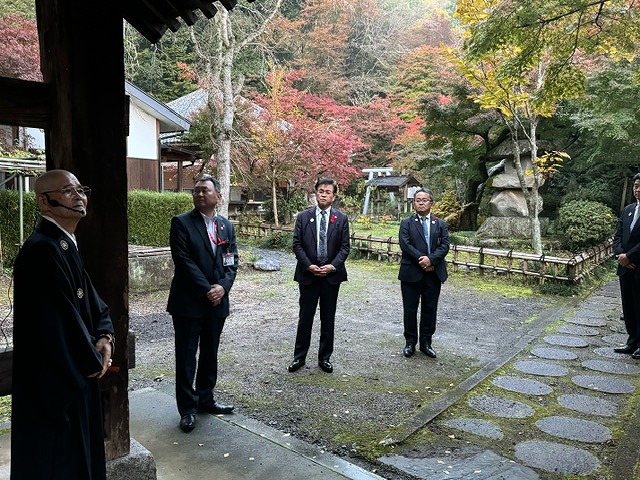 赤く色づいた木々に囲まれた石畳の庭に立った高橋市長と数人の男性が、袈裟を着た男性の話を聞いている様子の写真