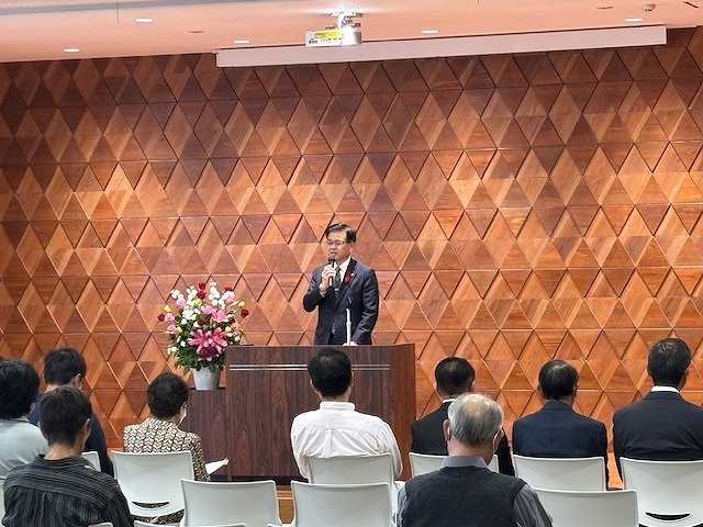 菱形の木目調の壁を背景に、演台に立った高橋市長が、客席に向かって話をしている写真