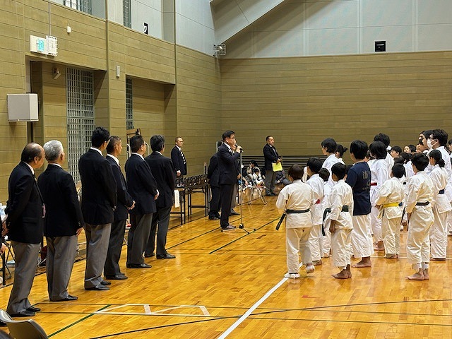 体育館に正装した複数の男性と空手着を着たたくさんの子どもたちが向かい合って立ち、子どもたちの前で高橋市長が話をしている様子の写真