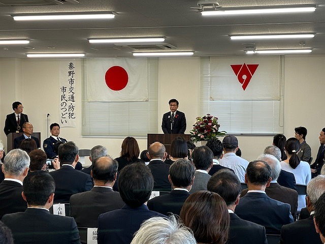 日本国旗と秦野市の市章が掲げられた壁の前に置かれた演台に立った高橋市長が、着席したたくさんの参加者に向かって話をしている写真