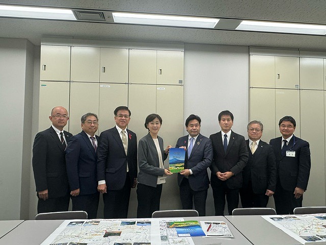 白いキャビネットが並んだ壁の前で、パンフレットを持った中央の男女を挟み、高橋市長を含む男性6人が並んで立っている記念写真