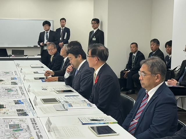 資料などを広げた長机の席に高橋市長を含む6人の男性が着席しており、その後ろには複数の男性たちがいる様子の写真