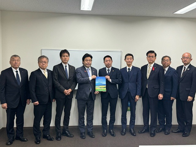 パンフレットを持った2人の男性を中心に、高橋市長を含む7人の男性が並んで立った記念写真