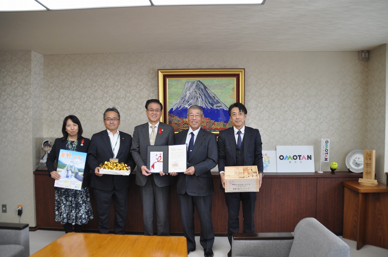 山の絵画が掛けられた壁を背に、男性と一緒に目録を持った高橋市長を中心に、ポスターを持った女性、たくさんの金色の鈴が入った箱を持った男性、木箱のようなものを持った男性が並んで立っている記念写真