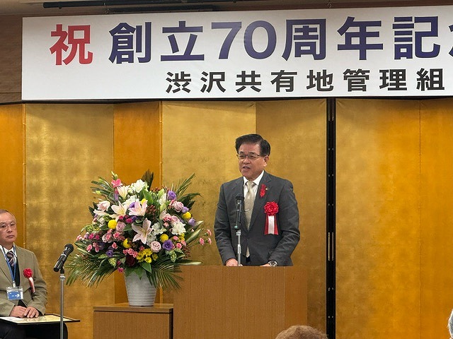創立70周年記念を祝う横断幕が掲げられた金屏風の前に、豪華な花を飾った演台が置かれ、そこに立った高橋市長がスピーチをしている様子の写真